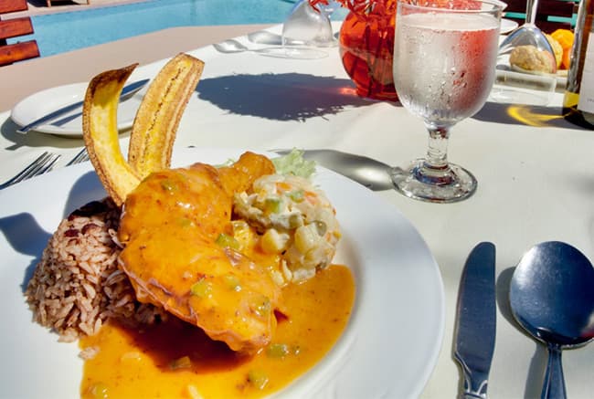 A plate of rice and chicken topped with a creamy sauce, accompanied by fried plantains and a side of coleslaw, with a glass of water nearby.