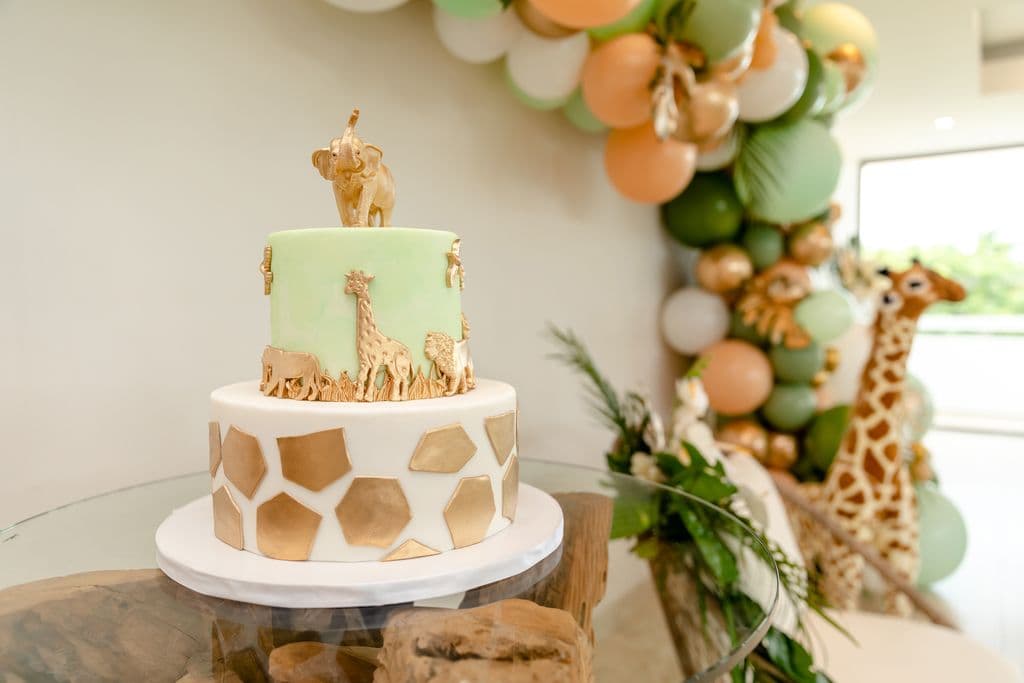 A two-tiered baby shower cake decorated with gold animal figurines, including a giraffe, elephant, and zebra. The bottom tier is covered in gold honeycomb-shaped patterns. A balloon arch and a plush giraffe toy are visible in the background.