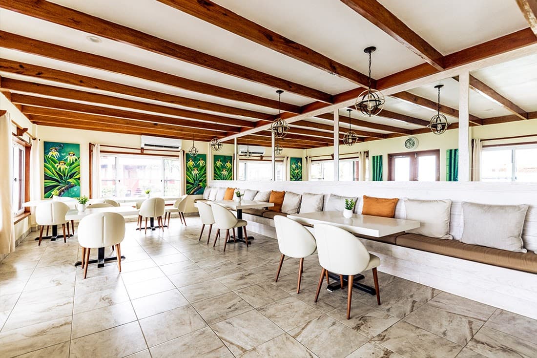 A spacious and modern restaurant with a high ceiling and exposed wooden beams. White tables and chairs are arranged throughout the room, with a cushioned bench seating area along one side.