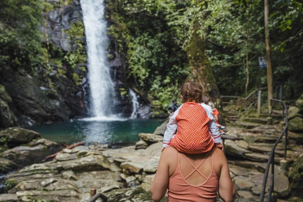 A woman carrying a toddler on her shoulders looks at a waterfall cascading down into a pool surrounded by rocky outcrops.