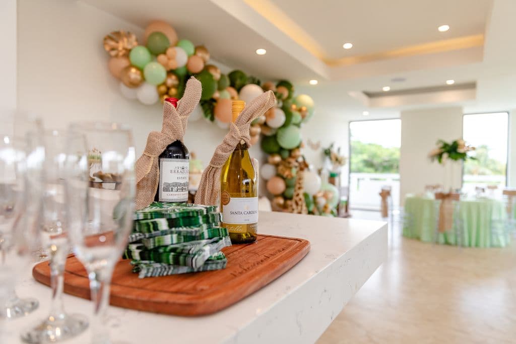 Two wine bottles wrapped in burlap are displayed on a wooden cutting board. Champagne flutes and a stack of green napkins are also on the board. A balloon garland and a decorated cake are visible in the background.