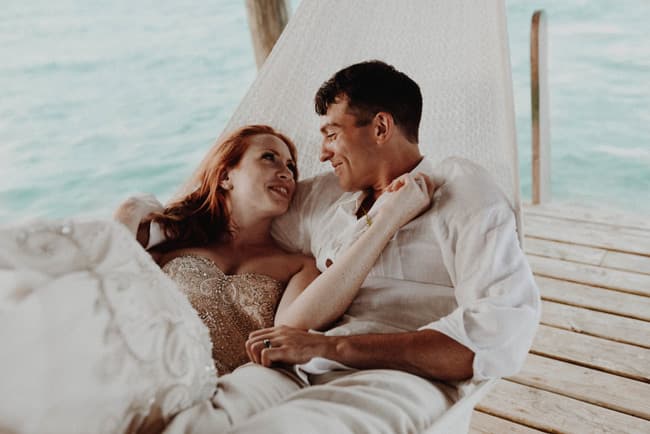A couple in wedding attire shares an intimate moment while lounging in a hammock by the water.