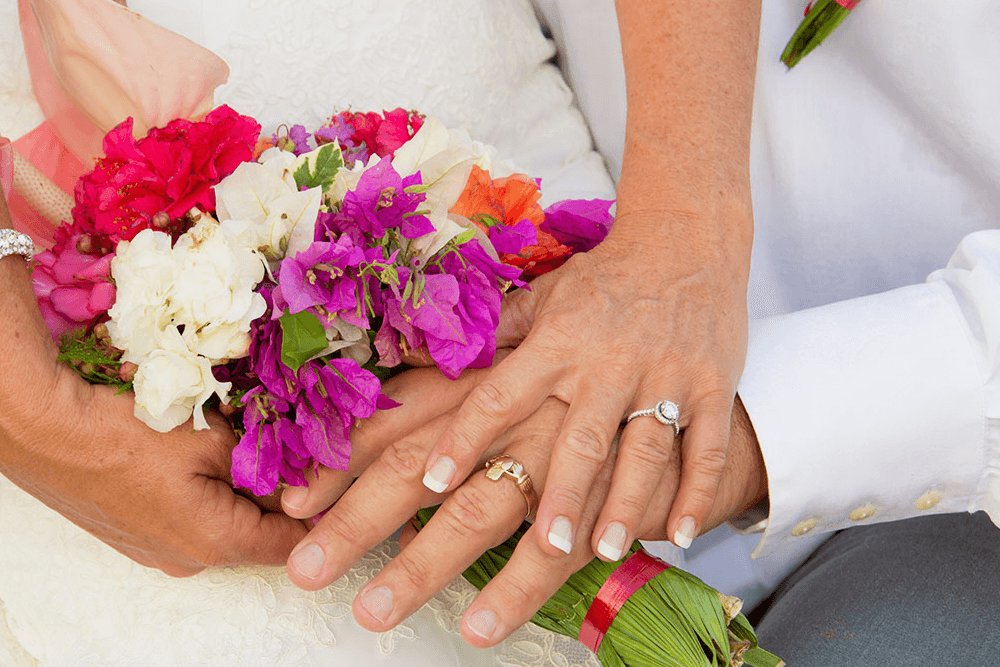 A bride and groom's hands holding a vibrant bouquet of pink, white, and red flowers.