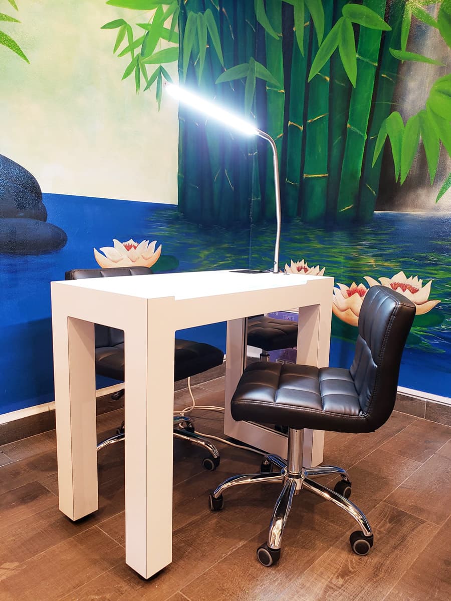 The image shows a nail salon station. There is a white table with a built-in LED lamp, two black swivel chairs, and a mural depicting bamboo, water, and lily pads in the background. The overall atmosphere appears clean and modern.