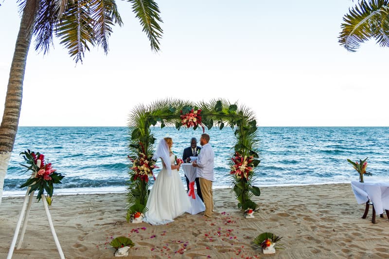 A couple exchanges vows underneath a floral arch on a beach.