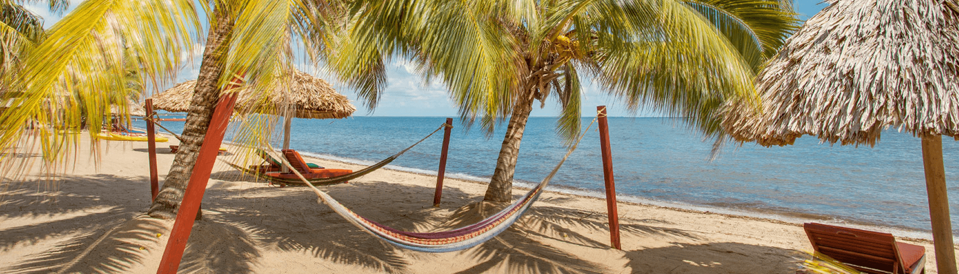 A serene beach scene with hammocks strung between palm trees. Umbrellas made of thatch provide shade on the sandy shore, and the calm turquoise water stretches out to the horizon.
