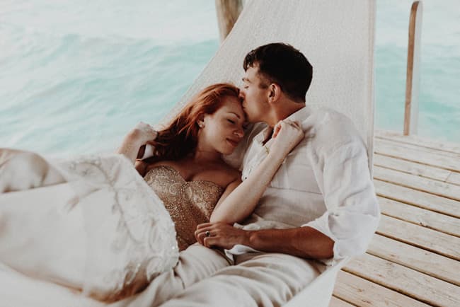 A couple relaxes together on a hammock by the water, sharing a tender moment.
