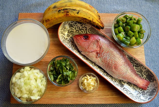 A selection of ingredients including a whole red fish, green okra, a banana, diced onion, chopped cilantro, minced garlic, and coconut milk on a wooden cutting board.