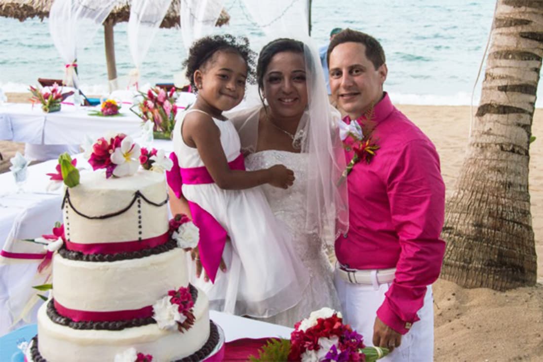 Bride, groom and flower girl in white and pink pose in front of a large wedding cake in front of a wedding arch on the beach.