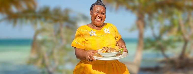 A woman in a vibrant yellow dress smiles while holding a plate of food against a tropical seaside backdrop. A woman in a vibrant yellow dress smiles while holding a plate of food against a tropical seaside backdrop.