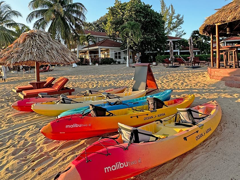 Colorful kayaks lined up on a sandy beach with palm trees and a thatched-roof hut in the background. Colorful kayaks lined up on a sandy beach with palm trees and a thatched-roof hut in the background.