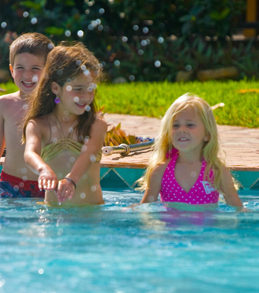 Three children play joyfully in a swimming pool, surrounded by bubbles and bright sunlight.