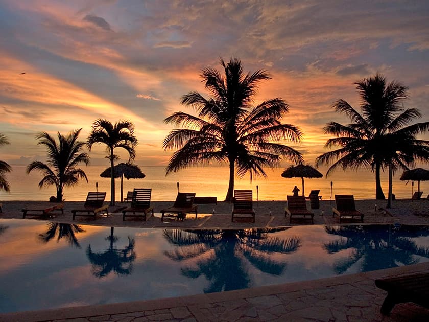 Sunset over a tranquil beach with palm trees and loungers reflected in the water.