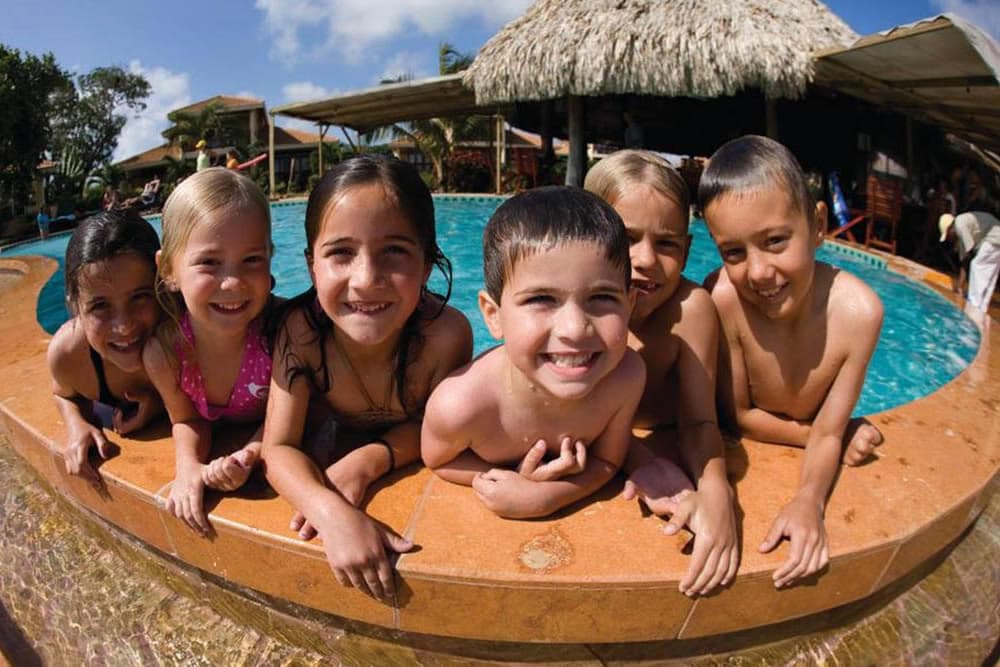 A group of six children smile toothily from the edge of a blue swimming pool on a sunny day.