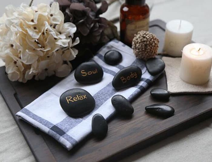 The image shows a spa-like setting with black stones arranged on a towel. The stones have the words "Soul," "Body," and "Relax" written on them. There are also candles, a flower arrangement, and a small bottle of essential oil in the background. The overall image suggests relaxation and rejuvenation.