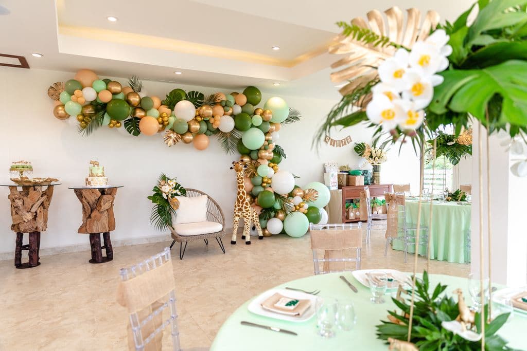 A beautifully decorated baby shower venue with a jungle theme. The room features a balloon arch in shades of green, gold, and peach. A plush giraffe toy sits in a chair near the arch. The tables are covered in light green tablecloths and adorned with floral centerpieces.