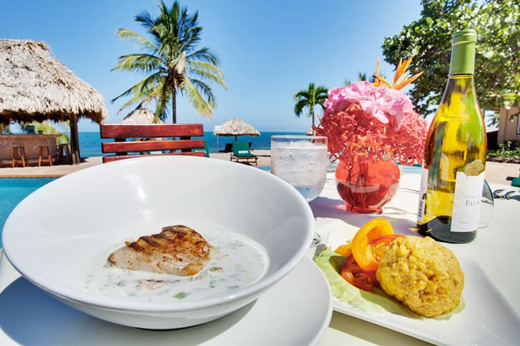 A beautifully plated fish dish with a side of cornbread and garnishes, set against a tropical backdrop with palm trees and a pool.