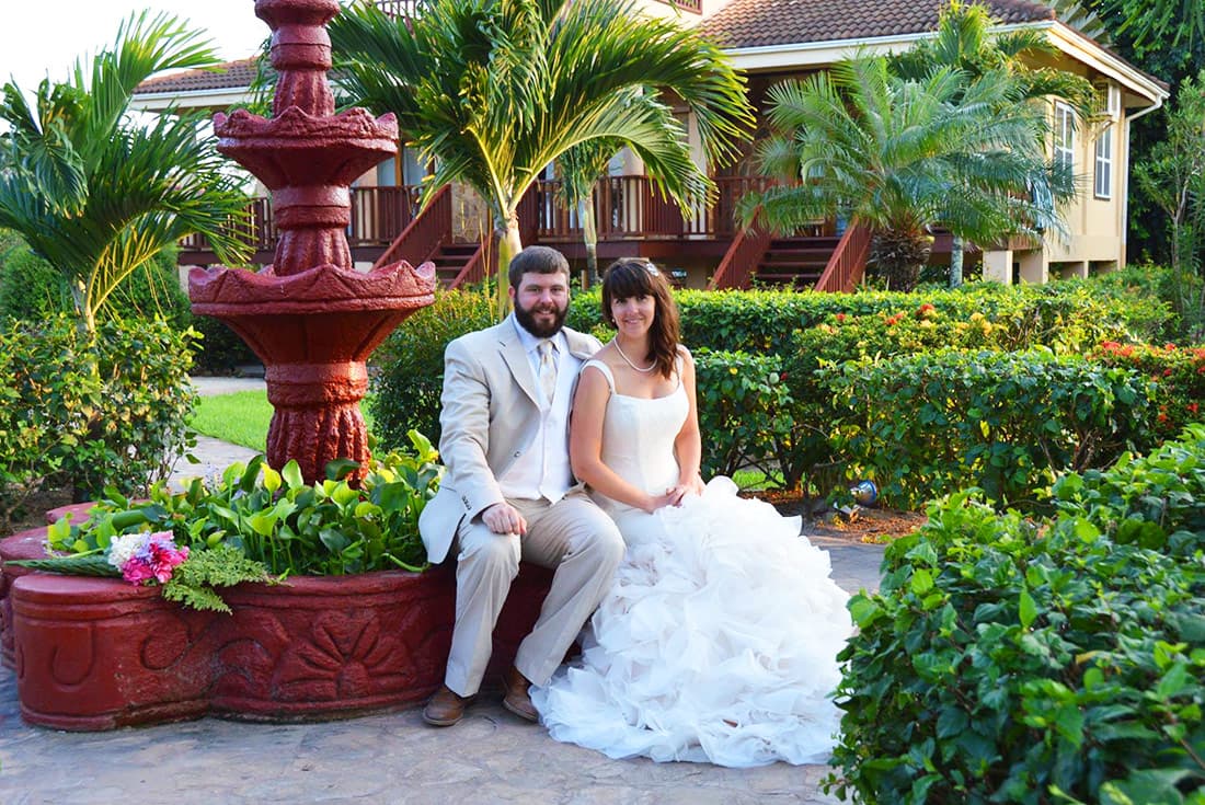A dark-haired bride and groom in wedding clothes pose happily on a fountain painted red, backed by beautiful villas and green foliage.