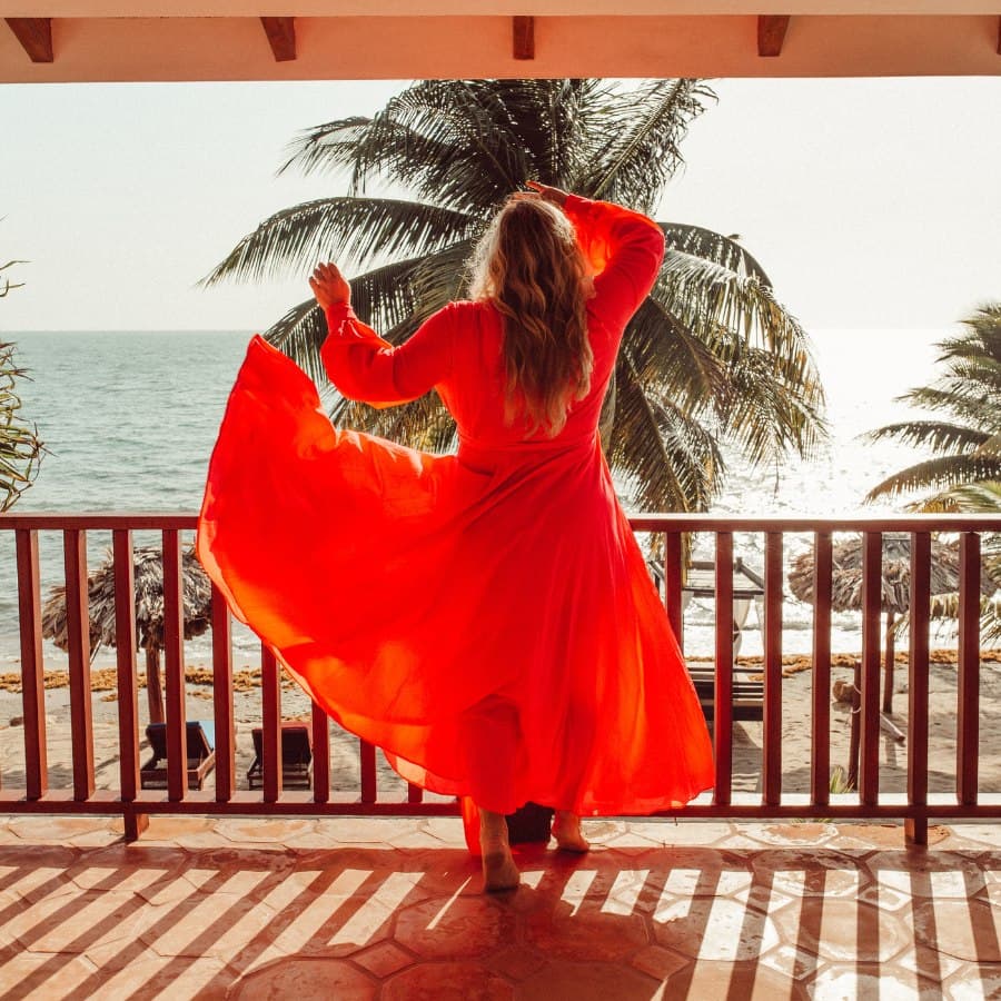 A person in a flowing red dress stands on a balcony, twirling against a backdrop of the ocean and palm trees.