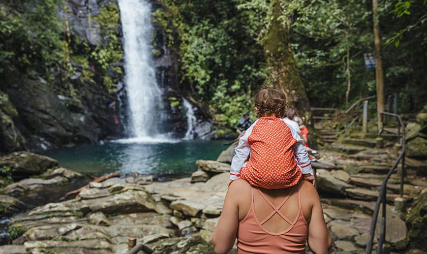 A person carries a child on their back while admiring a waterfall in a lush, green setting.