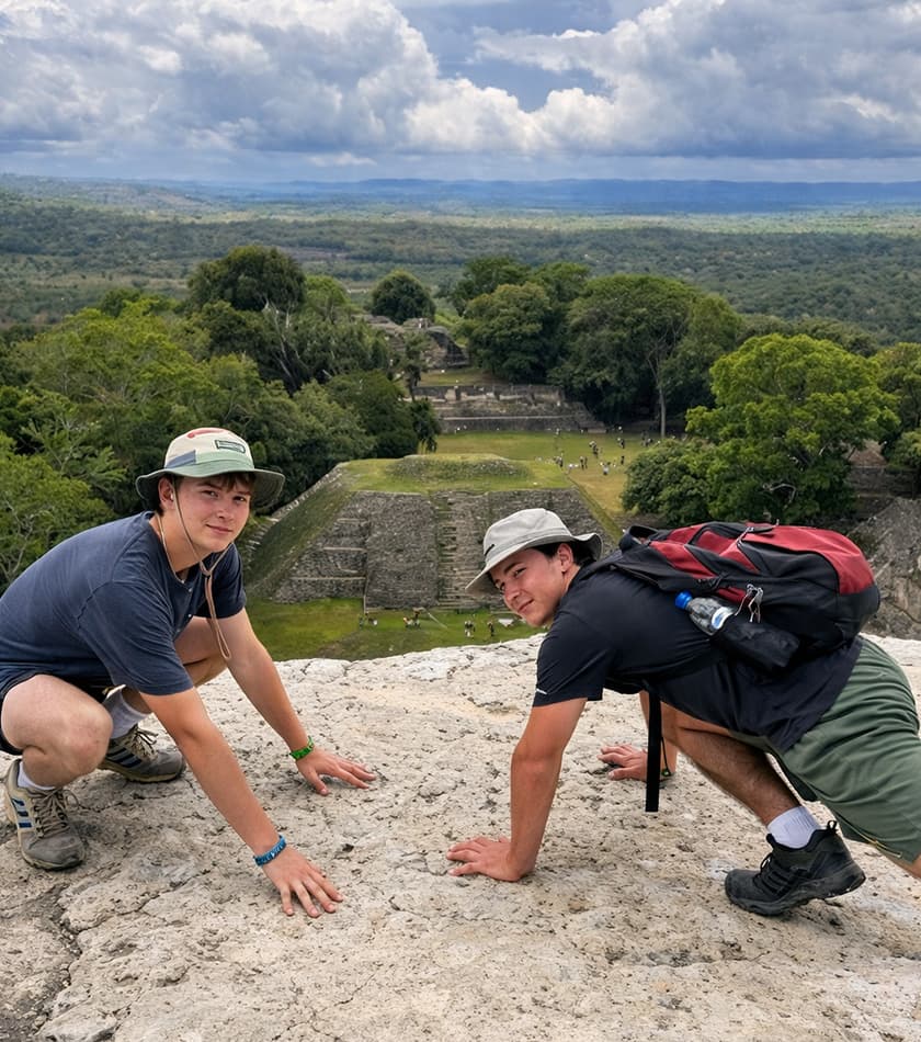 Two young men pose playfully on a rocky outcrop with ancient ruins and greenery in the background.