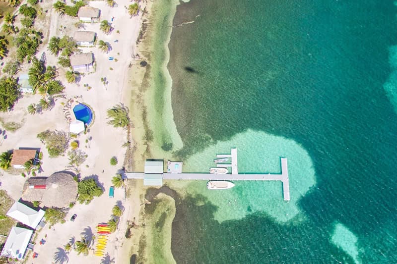 Aerial view of a beach with a pier and boats, surrounded by clear turquoise waters and greenery.