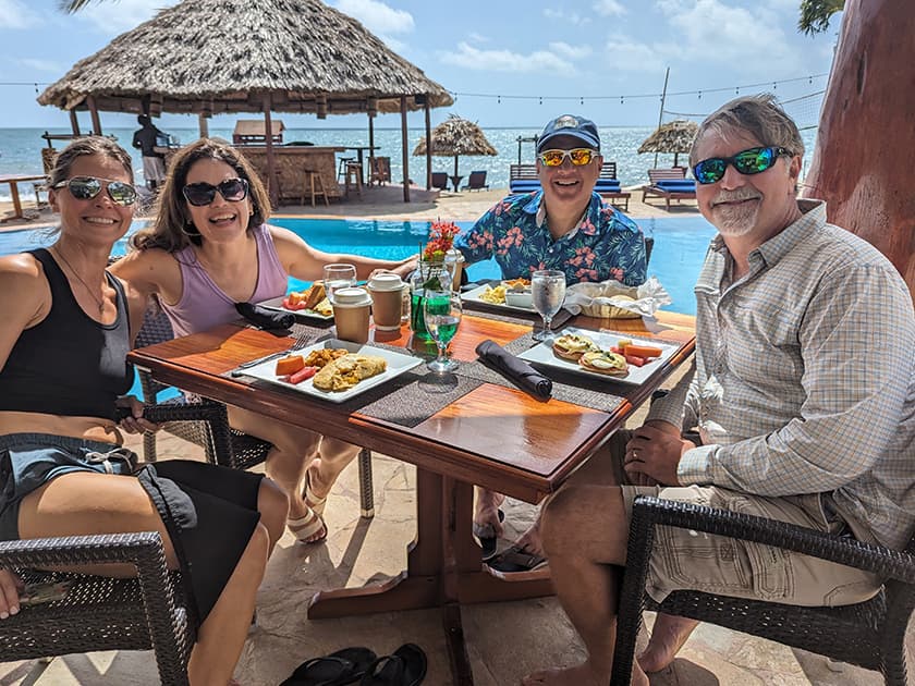 Four people enjoying a meal by a poolside in a sunny tropical setting. Four people enjoying a meal by a poolside in a sunny tropical setting.