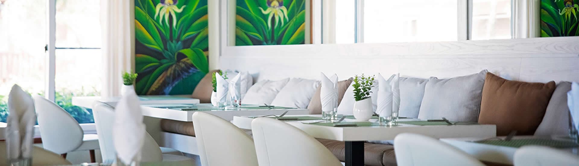 A modern restaurant interior with white walls and large windows. The restaurant features white tables and chairs, plush white and brown cushions, and a decorative wall mural of tropical plants.