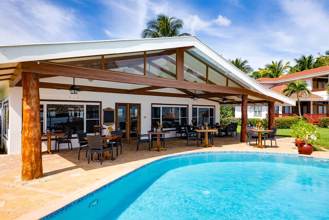 A beachfront restaurant with a covered outdoor seating area. The restaurant has a white facade and a wooden roof, with large windows overlooking the pool and ocean.