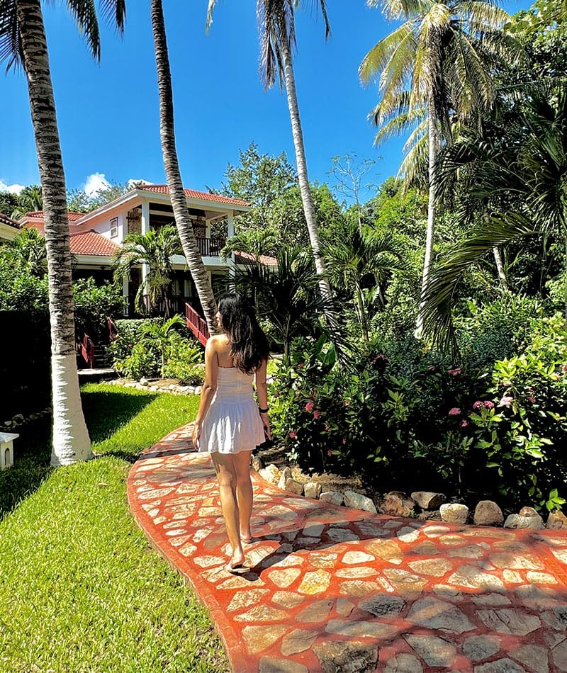 A woman in a light dress walks along a curved stone path surrounded by lush greenery and palm trees.