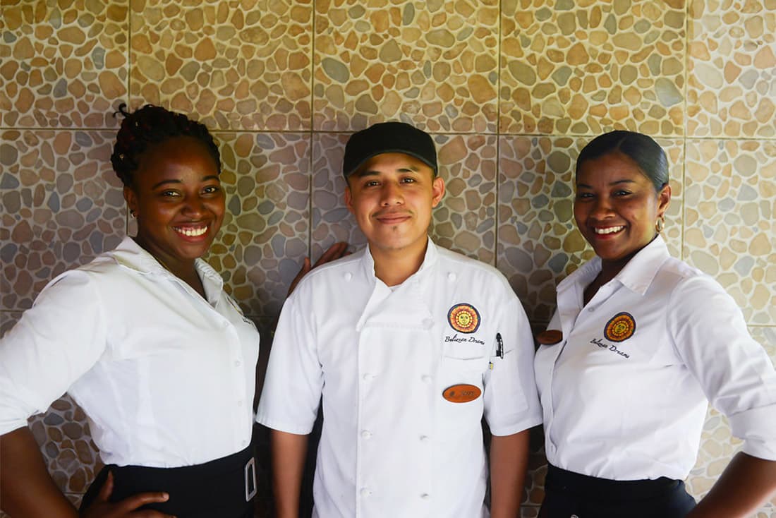 Three smiling restaurants in white tops stand in front of a yellow patterned wall.