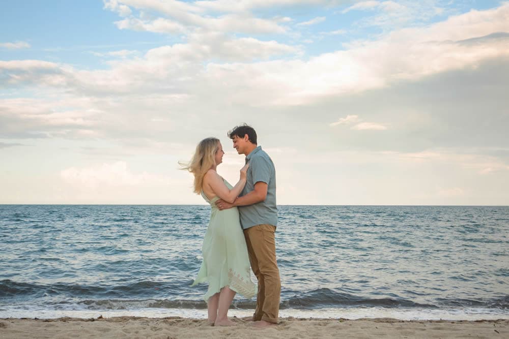 Man and woman holding each other on the beach with ocean and sky in background.