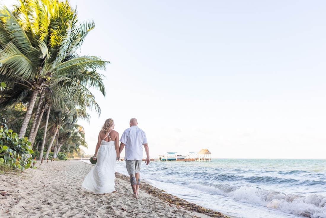 A blond bride in a gown and a bald groom with tattoos walk down the beach hand-in-hand.