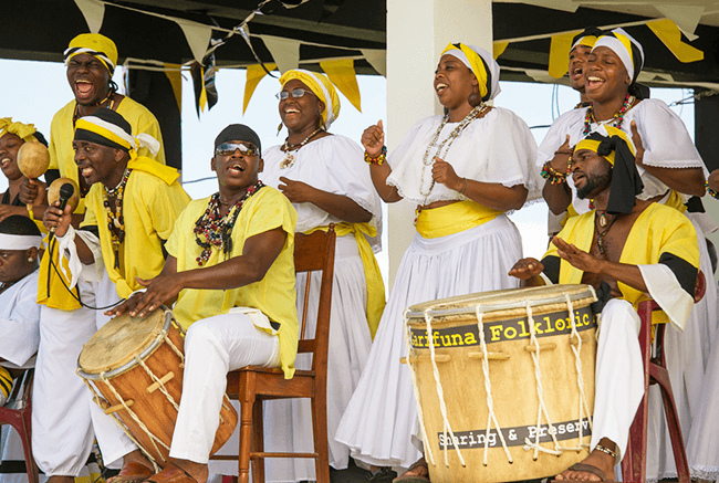 A lively group of performers in yellow and white attire play drums and sing together on stage.