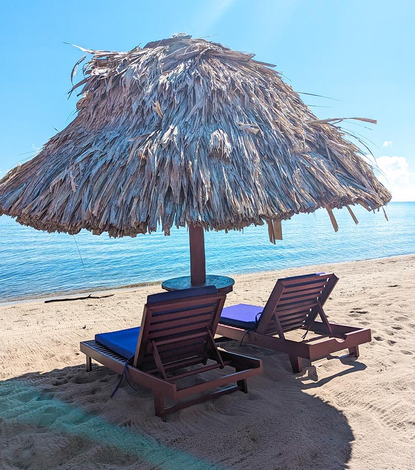 Two lounge chairs under a thatched umbrella on a sandy beach by the sea.