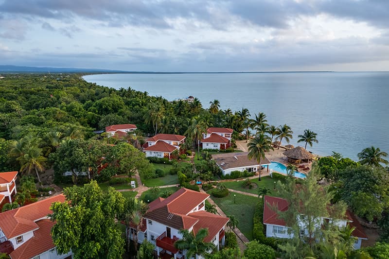 Aerial view of a tropical resort along the coast, featuring lush greenery, cottages, and a swimming pool near the ocean. Aerial view of a tropical resort along the coast, featuring lush greenery, cottages, and a swimming pool near the ocean.