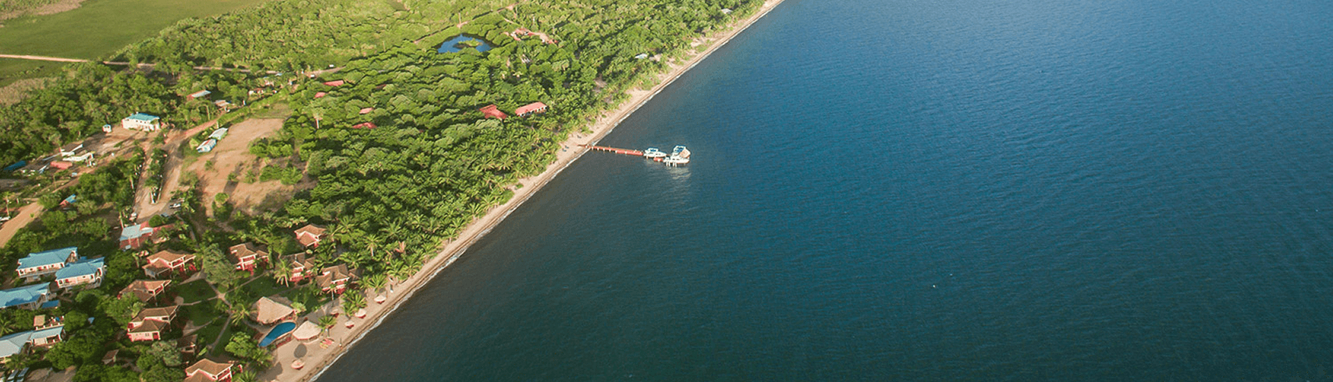 An aerial view of a secluded beach resort nestled along a coastline. The resort features a pier extending into the calm turquoise water, several small buildings surrounded by lush greenery, and a pool. The surrounding landscape includes dense forest and a small pond.