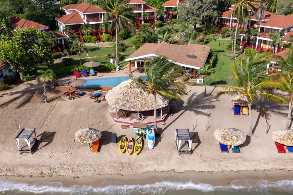 An aerial view of a tropical beach resort. The resort features multiple buildings with red roofs, a swimming pool, and a thatched-roof bar on the beach. Kayaks are lined up on the shore, and beach umbrellas provide shade for lounge chairs.