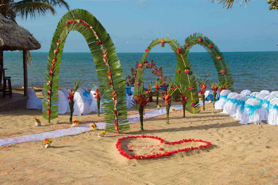 A beautiful beach wedding setup with three palm leaf archways decorated with colorful flowers. The ceremony area is adorned with white chairs and a heart-shaped design made of red flower petals on the sand. The ocean and a tropical beach backdrop complete the scene.
