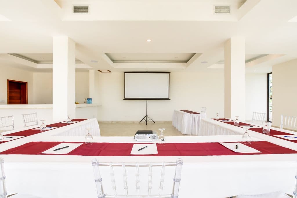 A modern meeting room with a U-shaped table arrangement. The room has a white tiled floor, white walls, and a high ceiling with exposed beams. A large projection screen and projector are at the front of the room. The table is covered in white tablecloths with red runners. Clear plastic chairs are arranged around the table. There is a bar area with counter stools in the background.