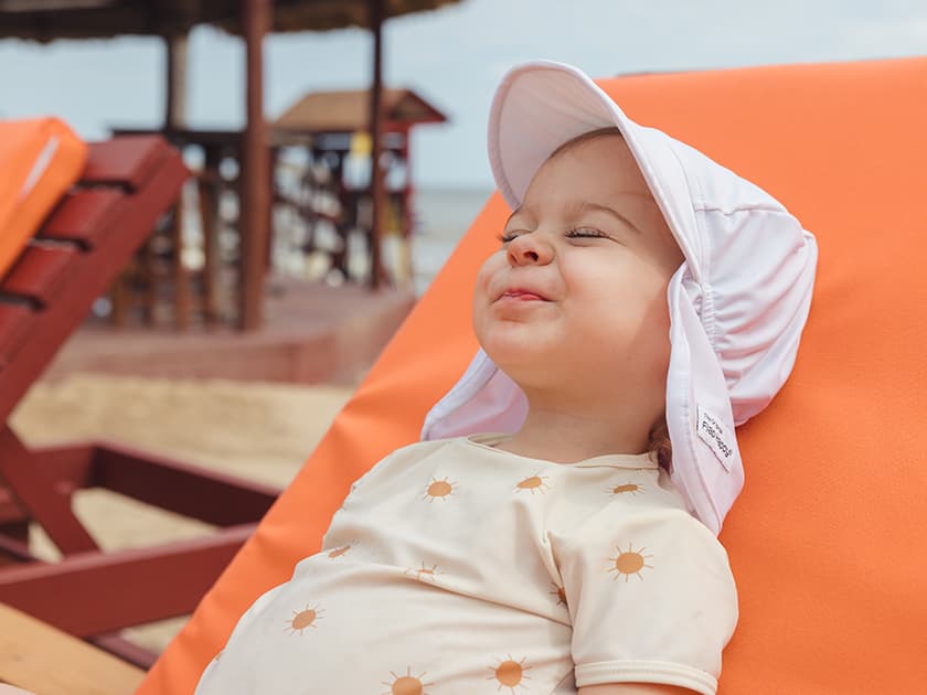 A smiling toddler lounges on a beach chair, wearing a sun hat and a shirt with sun patterns.